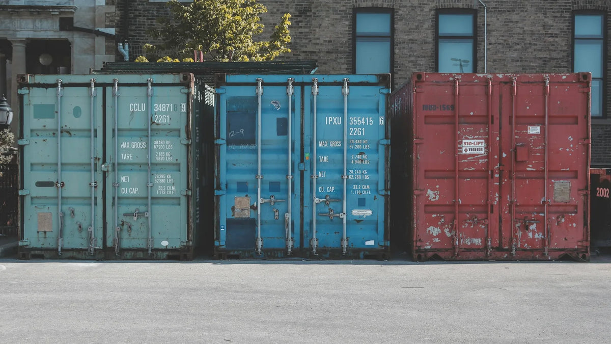Three used shipping containers in teal, blue and red parked side by side