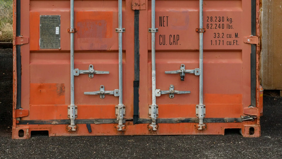 Orange shipping container standing level on a compacted gravel surface, doors closed