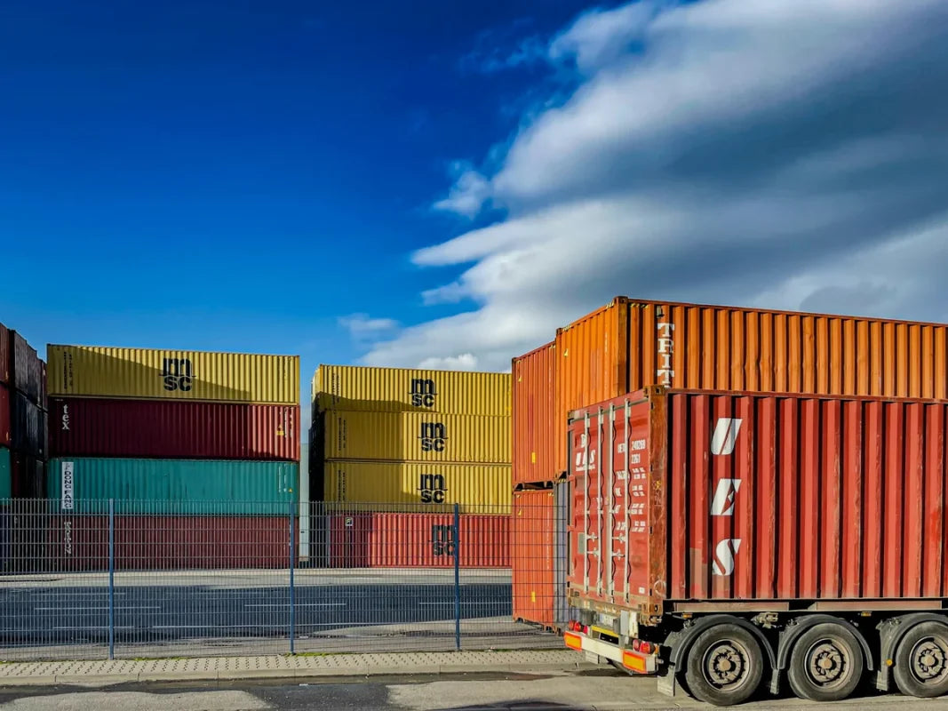 Truck trailer carrying a red shipping container past stacked containers in a yard